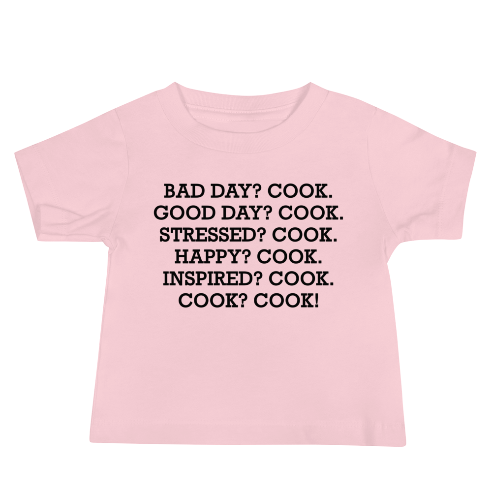 "Bad Day? Cook. Good Day? Cook. Stressed? Cook. Happy? Cook. Inspired? Cook. Cook? Cook!" printed on a pink baby jersey short sleeve tee laying on a flat surface