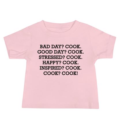 "Bad Day? Cook. Good Day? Cook. Stressed? Cook. Happy? Cook. Inspired? Cook. Cook? Cook!" printed on a pink baby jersey short sleeve tee laying on a flat surface