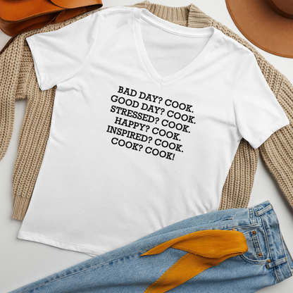 "Bad Day? Cook. Good Day? Cook. Stressed? Cook. Happy? Cook. Inspired? Cook. Cook? Cook!" printed on a solid white blend women's relaxed v-neck t-shirt laying on a flat surface amongst other clothes