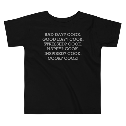 "Bad Day? Cook. Good Day? Cook. Stressed? Cook. Happy? Cook. Inspired? Cook. Cook? Cook!" printed on a black toddler short sleeve tee laying on a flat surface