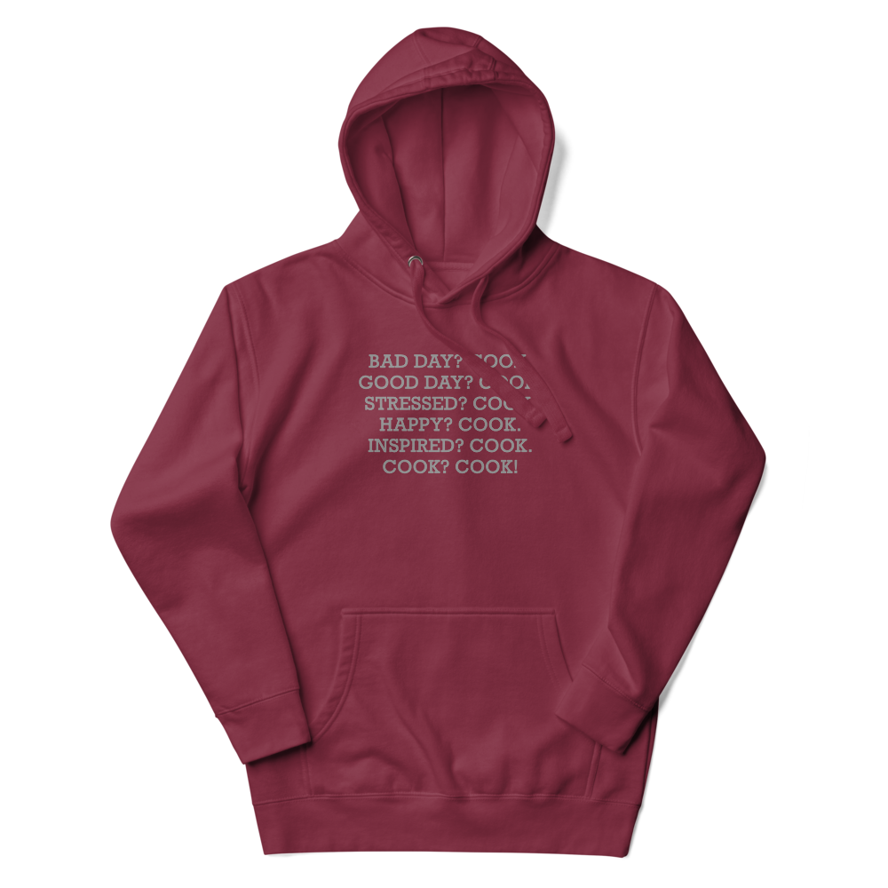 "Bad Day? Cook. Good Day? Cook. Stressed? Cook. Happy? Cook. Inspired? Cook. Cook? Cook!" printed on a maroon unisex hoodie laying on a flat surface with the hood up