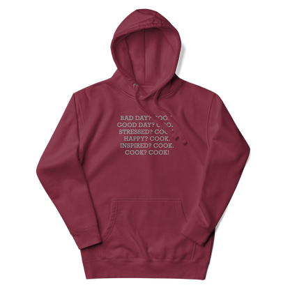 "Bad Day? Cook. Good Day? Cook. Stressed? Cook. Happy? Cook. Inspired? Cook. Cook? Cook!" printed on a maroon unisex hoodie laying on a flat surface with the hood up