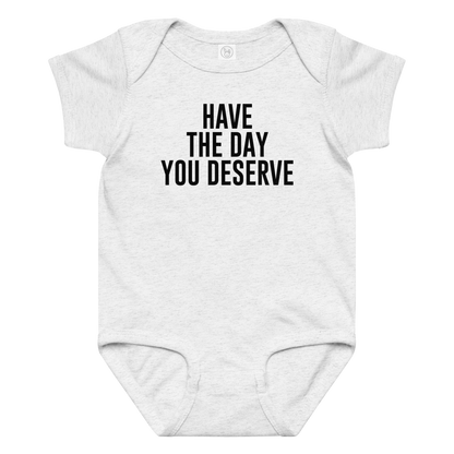 "Have the day you deserve" printed on a ash baby jersey bodysuit laying on a flat surface amongst other clothes
