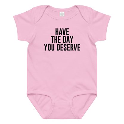 "Have the day you deserve" printed on a pink baby jersey bodysuit laying on a flat surface amongst other clothes