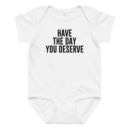 "Have the day you deserve" printed on a white baby jersey bodysuit laying on a flat surface amongst other clothes