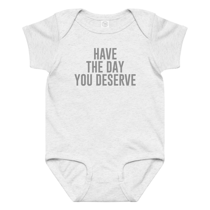 "Have the day you deserve" printed on a ash baby jersey bodysuit laying on a flat surface amongst other clothes