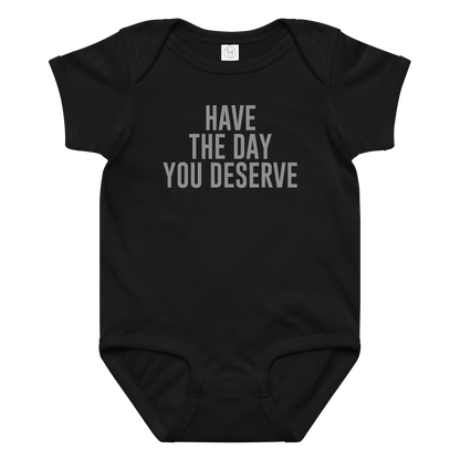 "Have the day you deserve" printed on a black baby jersey bodysuit laying on a flat surface amongst other clothes