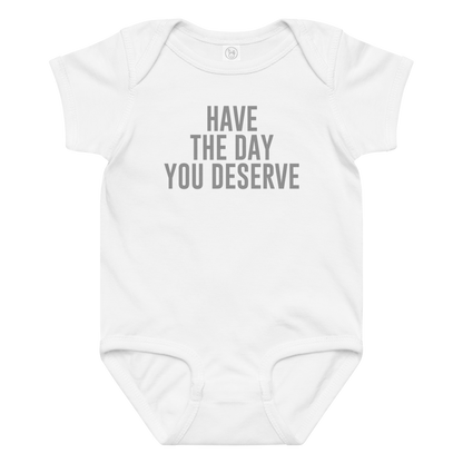 "Have the day you deserve" printed on a white baby jersey bodysuit laying on a flat surface amongst other clothes