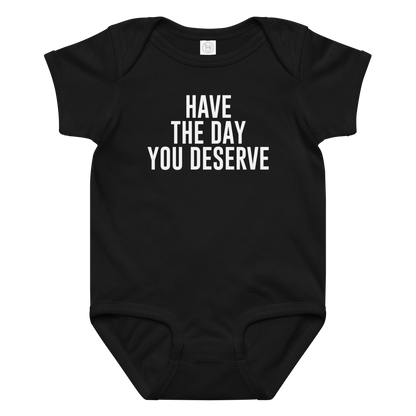 "Have the day you deserve" printed on a black baby jersey bodysuit laying on a flat surface amongst other clothes