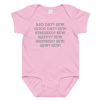 "Bad Day? Sew. Good Day? Sew. Stressed? Sew. Happy? Sew. Inspired? Sew. Sew? Sew!" printed on a pink baby jersey bodysuit laying on a flat surface amongst other clothes