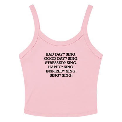 "Bad Day? Sing. Good Day? Sing. Stressed? Sing. Happy? Sing. Inspired? Sing. Sing? Sing!" printed on a solid pink blend women's micro-rib tanktop laying on a flat surface amongst sunglasses and shorts