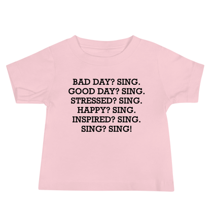 "Bad Day? Sing. Good Day? Sing. Stressed? Sing. Happy? Sing. Inspired? Sing. Sing? Sing!" printed on a pink baby jersey short sleeve tee laying on a flat surface