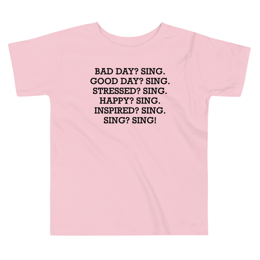 "Bad Day? Sing. Good Day? Sing. Stressed? Sing. Happy? Sing. Inspired? Sing. Sing? Sing!" printed on a pink toddler short sleeve tee laying on a flat surface