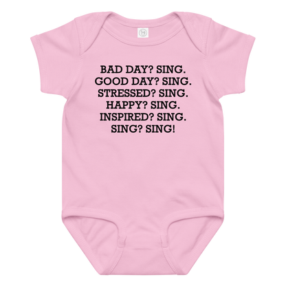 "Bad Day? Sing. Good Day? Sing. Stressed? Sing. Happy? Sing. Inspired? Sing. Sing? Sing!" printed on a pink baby jersey bodysuit laying on a flat surface amongst other clothes