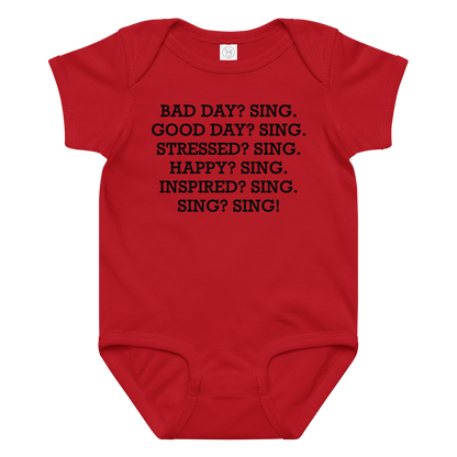 "Bad Day? Sing. Good Day? Sing. Stressed? Sing. Happy? Sing. Inspired? Sing. Sing? Sing!" printed on a red baby jersey bodysuit laying on a flat surface amongst other clothes