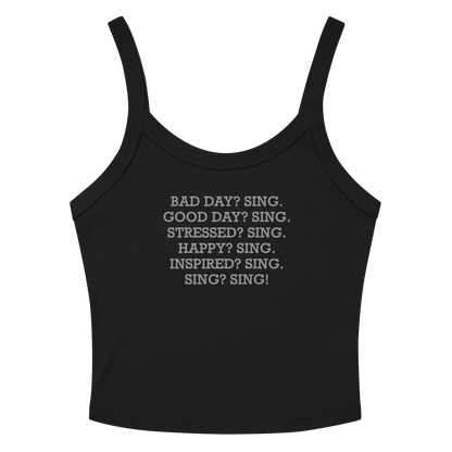 "Bad Day? Sing. Good Day? Sing. Stressed? Sing. Happy? Sing. Inspired? Sing. Sing? Sing!" printed on a solid black blend women's micro-rib tanktop laying on a flat surface amongst sunglasses and shorts