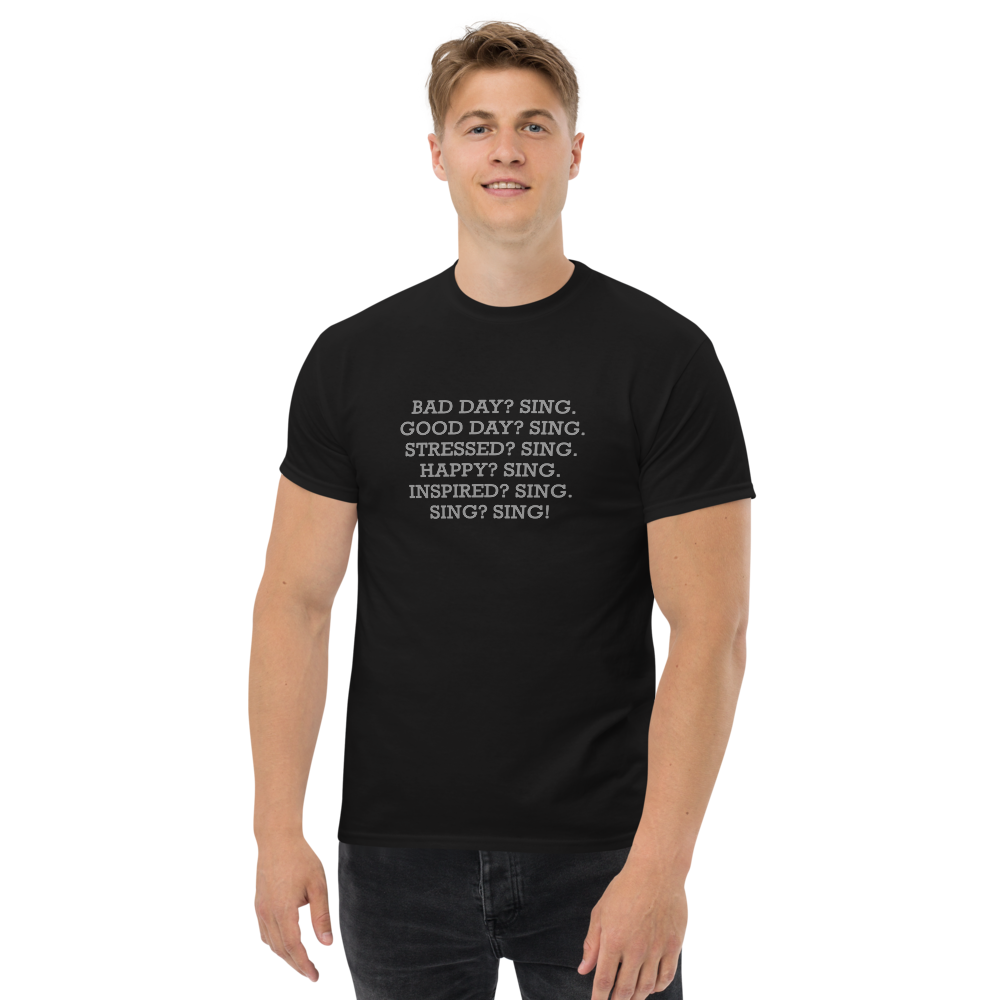 "Bad Day? Sing. Good Day? Sing. Stressed? Sing. Happy? Sing. Inspired? Sing. Sing? Sing!" printed on a black unisex classic tee worn by a male