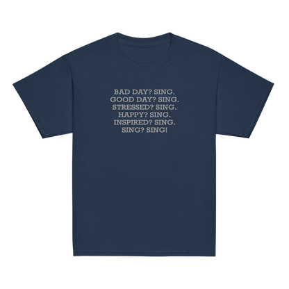 "Bad Day? Sing. Good Day? Sing. Stressed? Sing. Happy? Sing. Inspired? Sing. Sing? Sing!" printed on a navy youth classic tee laying on a flat surface