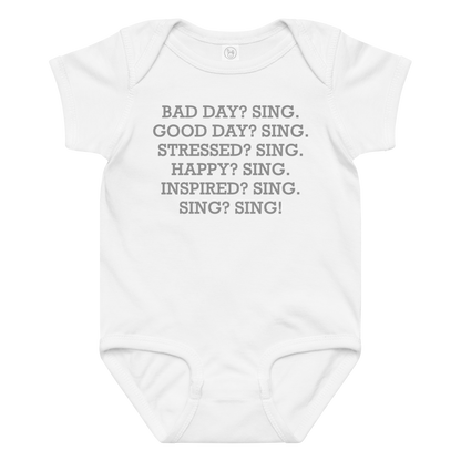 "Bad Day? Sing. Good Day? Sing. Stressed? Sing. Happy? Sing. Inspired? Sing. Sing? Sing!" printed on a white baby jersey bodysuit laying on a flat surface amongst other clothes