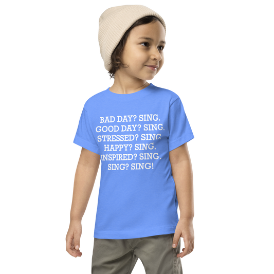 "Bad Day? Sing. Good Day? Sing. Stressed? Sing. Happy? Sing. Inspired? Sing. Sing? Sing!" printed on a heather columbia blue toddler short sleeve tee worn by a male wearing a cap