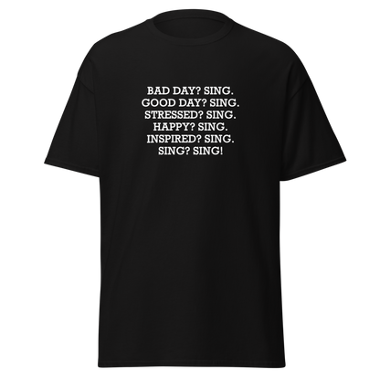 "Bad Day? Sing. Good Day? Sing. Stressed? Sing. Happy? Sing. Inspired? Sing. Sing? Sing!" printed on a black unisex classic tee laying on a flat surface