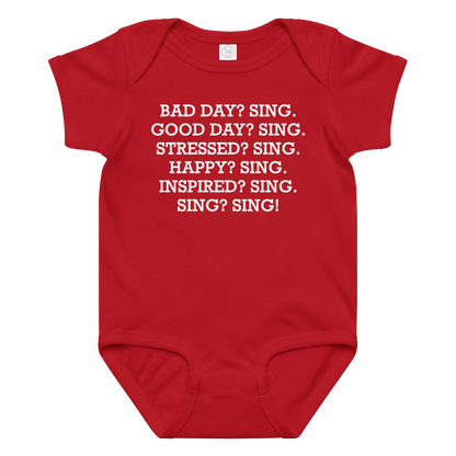 "Bad Day? Sing. Good Day? Sing. Stressed? Sing. Happy? Sing. Inspired? Sing. Sing? Sing!" printed on a red baby jersey bodysuit laying on a flat surface amongst other clothes