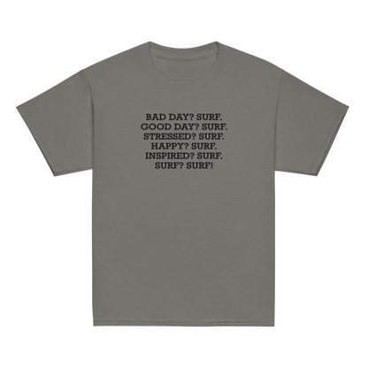 "Bad Day? Surf. Good Day? Surf. Stressed? Surf. Happy? Surf. Inspired? Surf. Surf? Surf!" printed on a charcoal youth classic tee laying on a flat surface