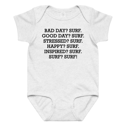 "Bad Day? Surf. Good Day? Surf. Stressed? Surf. Happy? Surf. Inspired? Surf. Surf? Surf!" printed on a ash baby jersey bodysuit laying on a flat surface amongst other clothes