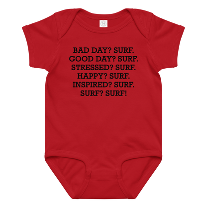 "Bad Day? Surf. Good Day? Surf. Stressed? Surf. Happy? Surf. Inspired? Surf. Surf? Surf!" printed on a red baby jersey bodysuit laying on a flat surface amongst other clothes