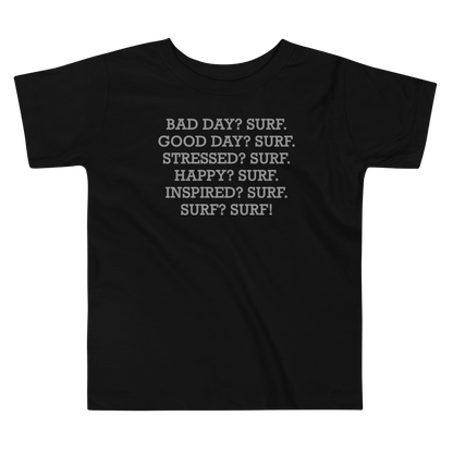 "Bad Day? Surf. Good Day? Surf. Stressed? Surf. Happy? Surf. Inspired? Surf. Surf? Surf!" printed on a black toddler short sleeve tee laying on a flat surface