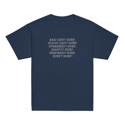 "Bad Day? Surf. Good Day? Surf. Stressed? Surf. Happy? Surf. Inspired? Surf. Surf? Surf!" printed on a navy youth classic tee laying on a flat surface