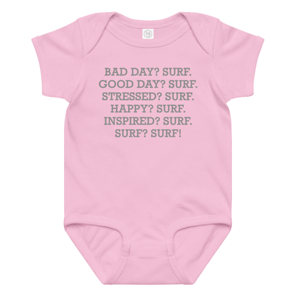 "Bad Day? Surf. Good Day? Surf. Stressed? Surf. Happy? Surf. Inspired? Surf. Surf? Surf!" printed on a pink baby jersey bodysuit laying on a flat surface amongst other clothes