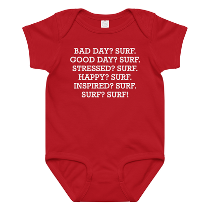 "Bad Day? Surf. Good Day? Surf. Stressed? Surf. Happy? Surf. Inspired? Surf. Surf? Surf!" printed on a red baby jersey bodysuit laying on a flat surface amongst other clothes