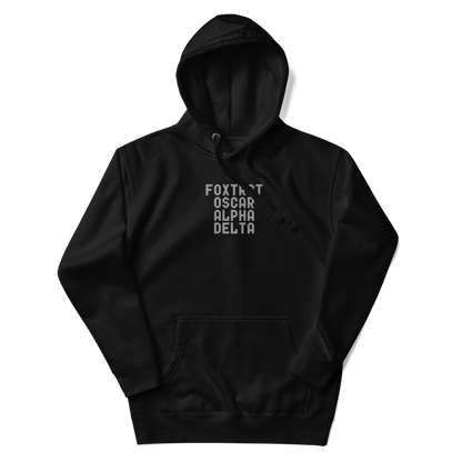 "Foxtrot Oscar Alpha Delta" printed on a black unisex hoodie laying on a flat surface with the hood up