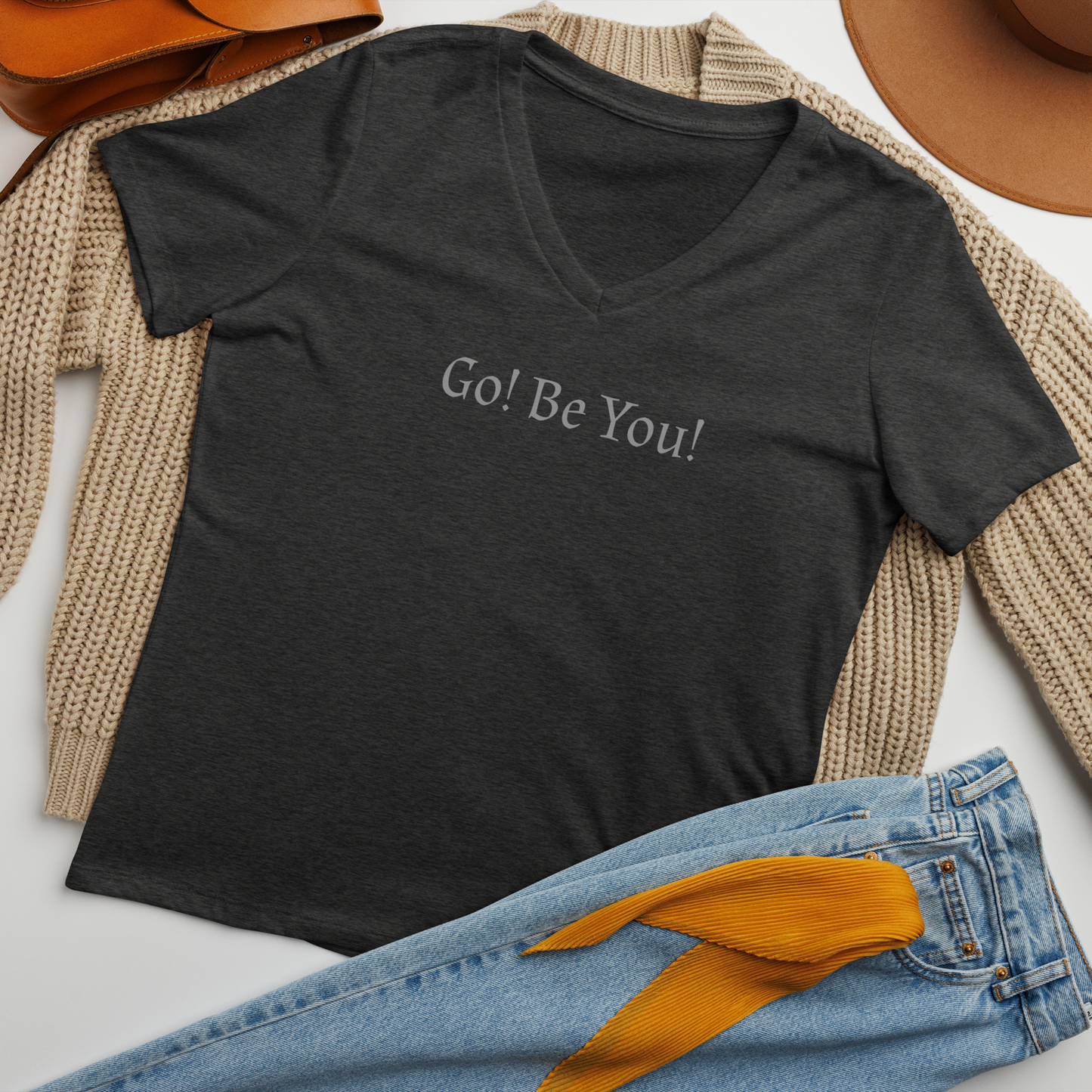 "Go! Be You!" printed on a dark grey heather women's relaxed v-neck t-shirt laying on a flat surface amongst other clothes