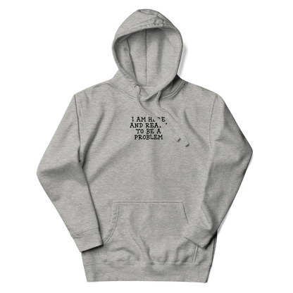 "I am here and ready to be a problem" printed on a carbon grey unisex hoodie laying on a flat surface with the hood up