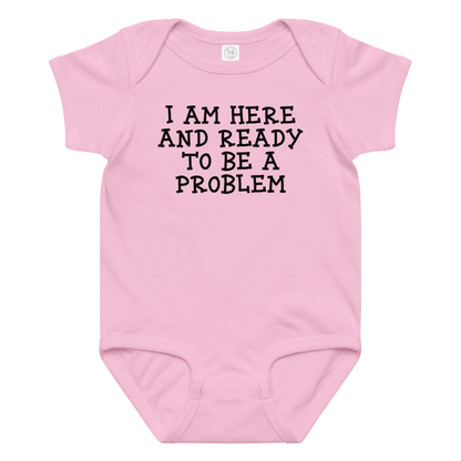 "I am here and ready to be a problem" printed on a pink baby jersey bodysuit laying on a flat surface amongst other clothes