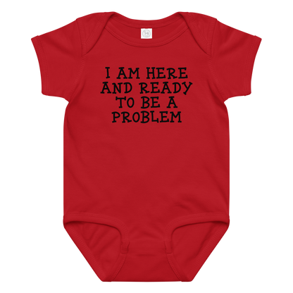 "I am here and ready to be a problem" printed on a red baby jersey bodysuit laying on a flat surface amongst other clothes