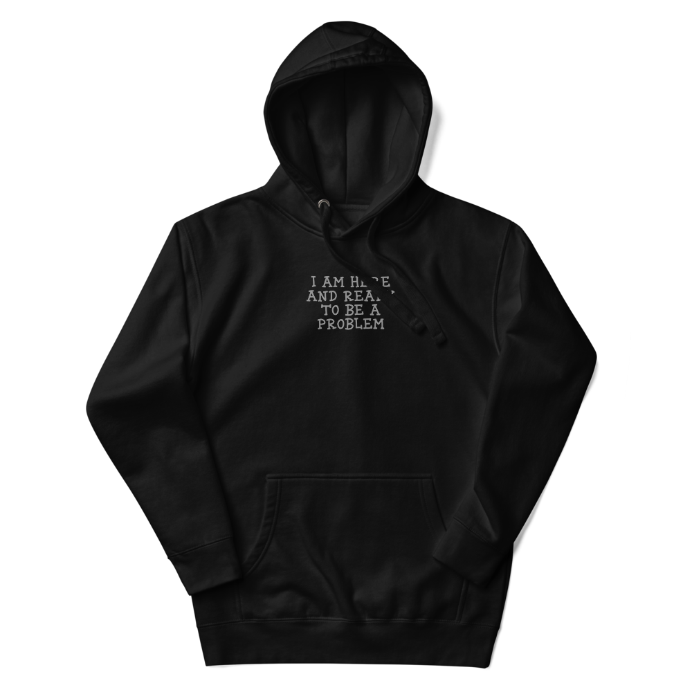 "I am here and ready to be a problem" printed on a black unisex hoodie laying on a flat surface with the hood up