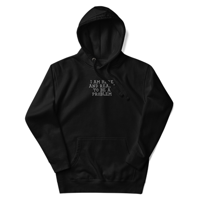 "I am here and ready to be a problem" printed on a black unisex hoodie laying on a flat surface with the hood up