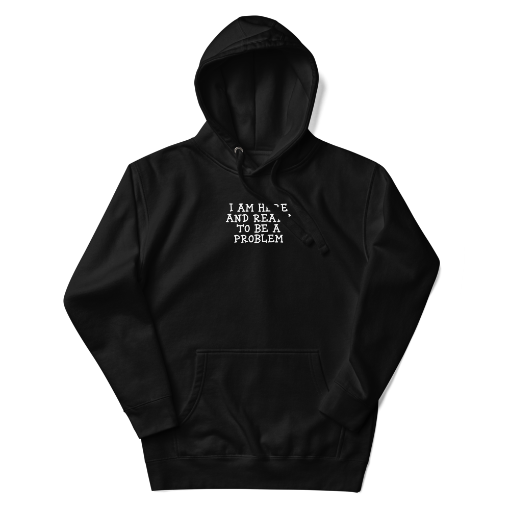 "I am here and ready to be a problem" printed on a black unisex hoodie laying on a flat surface with the hood up