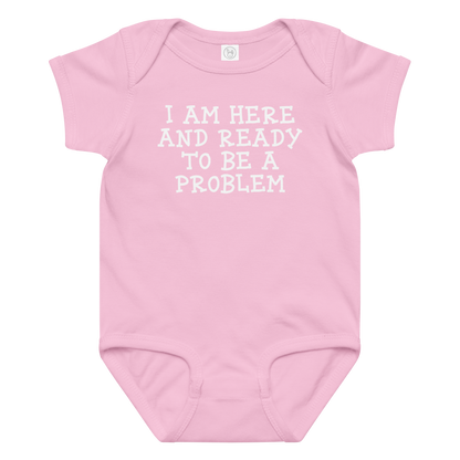"I am here and ready to be a problem" printed on a pink baby jersey bodysuit laying on a flat surface amongst other clothes