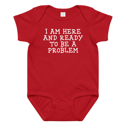 "I am here and ready to be a problem" printed on a red baby jersey bodysuit laying on a flat surface amongst other clothes