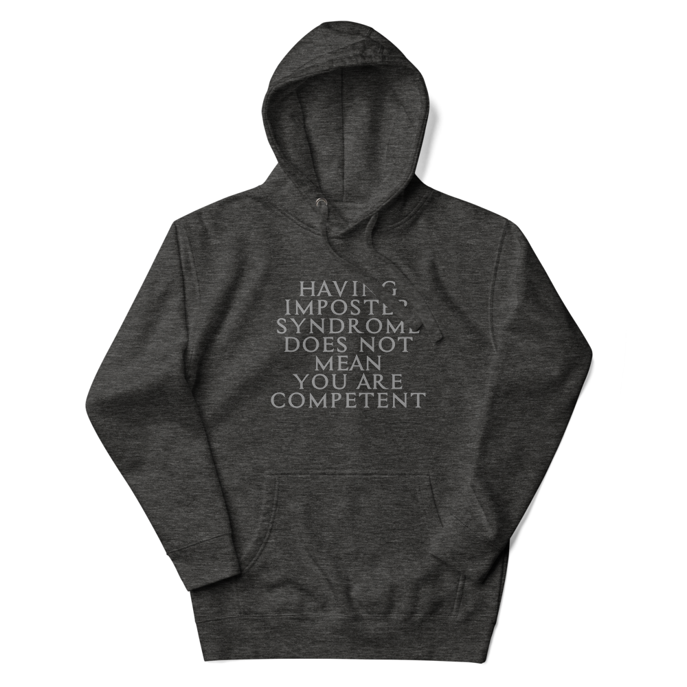 "Having Imposter Syndrome does not mean you are competent" printed on a charcoal heather unisex hoodie laying on a flat surface with the hood up