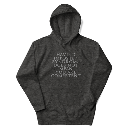 "Having Imposter Syndrome does not mean you are competent" printed on a charcoal heather unisex hoodie laying on a flat surface with the hood up