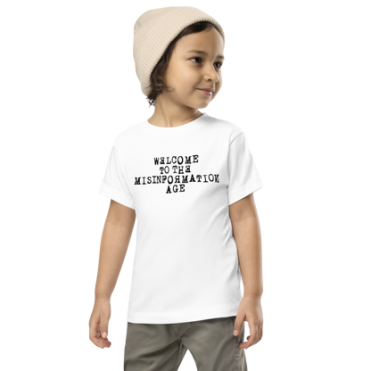 "Welcome to the misinformation age" printed on a white toddler short sleeve tee worn by a male wearing a cap