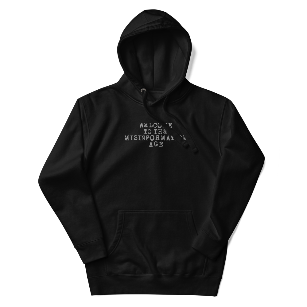 "Welcome to the misinformation age" printed on a black unisex hoodie laying on a flat surface with the hood up