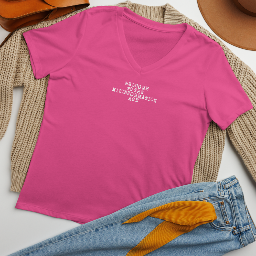 "Welcome to the misinformation age" printed on a berry women's relaxed v-neck t-shirt laying on a flat surface amongst other clothes