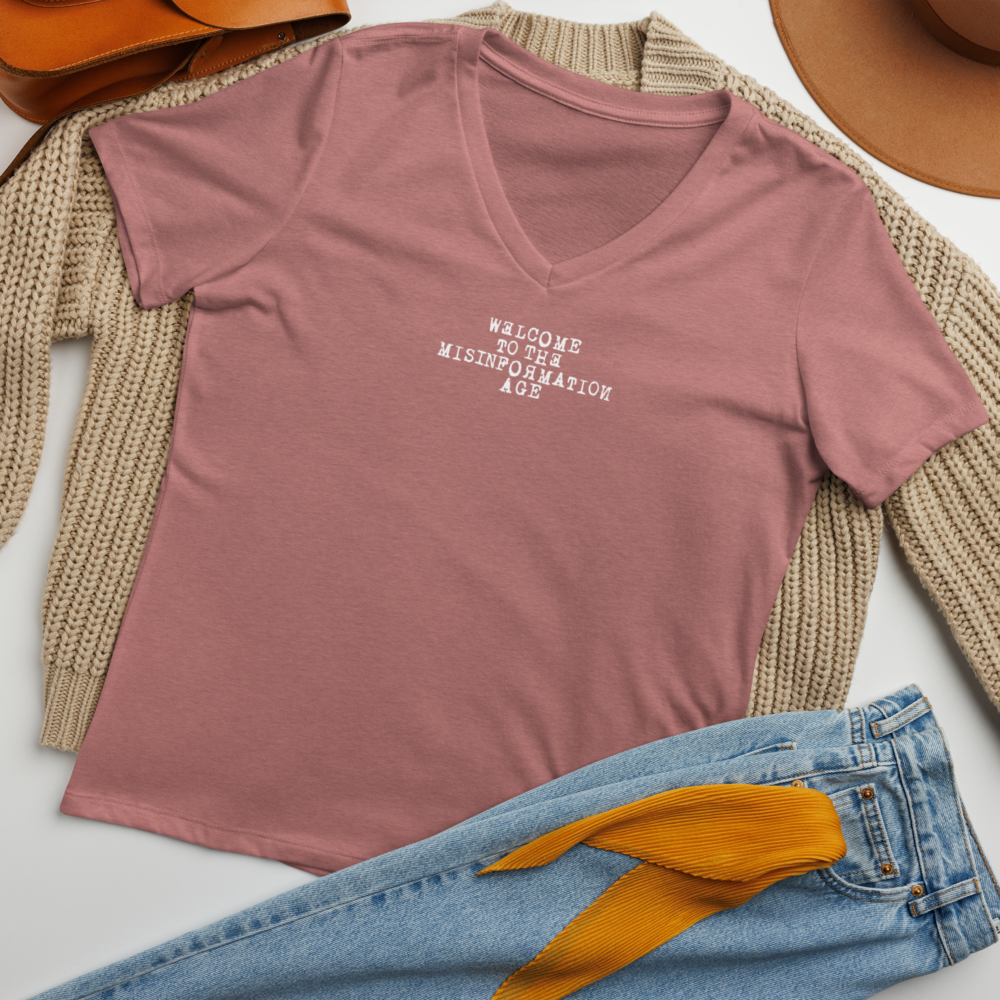 "Welcome to the misinformation age" printed on a heather mauve women's relaxed v-neck t-shirt laying on a flat surface amongst other clothes