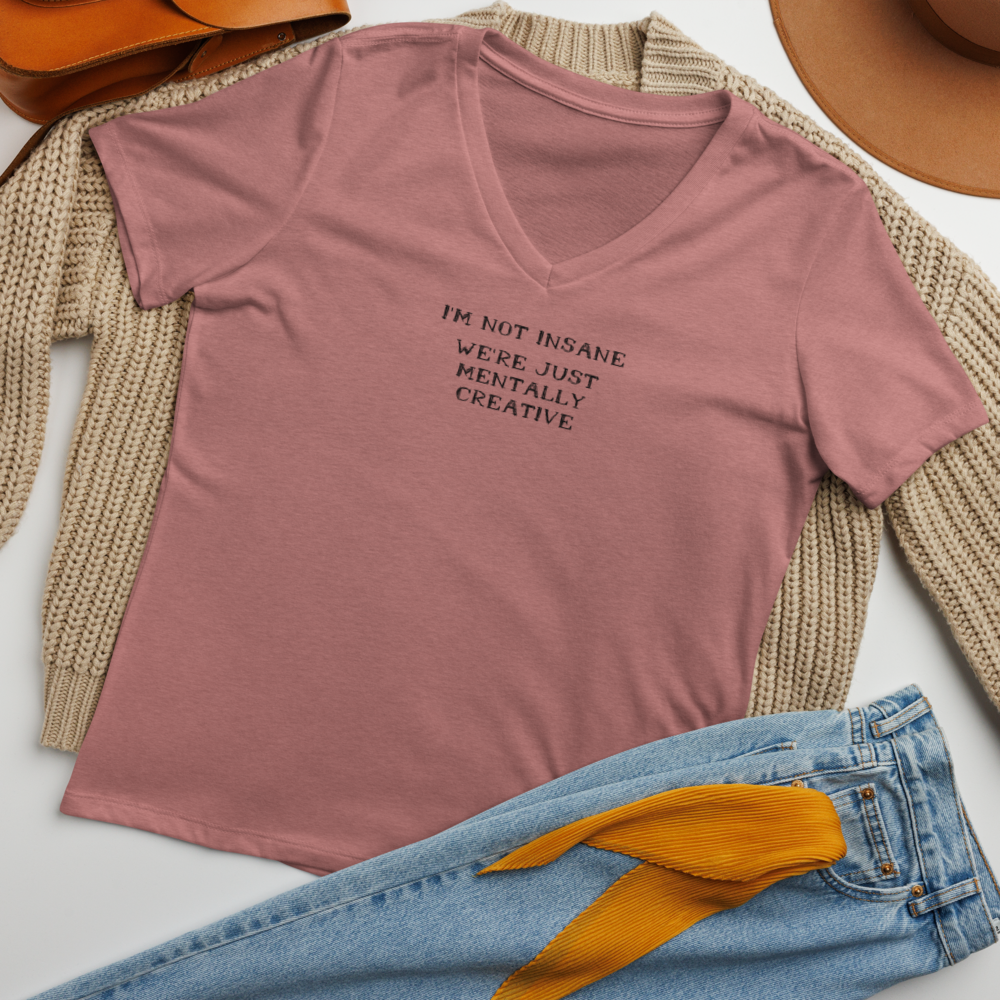 "I’m not insane   We’re just mentally creative" printed on a heather mauve women's relaxed v-neck t-shirt laying on a flat surface amongst other clothes